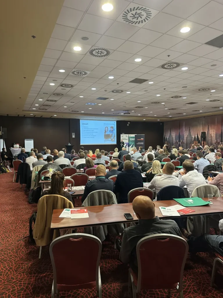 Konferenční sál plný účastníků sledujících prezentaci na plátně během odborné konference.